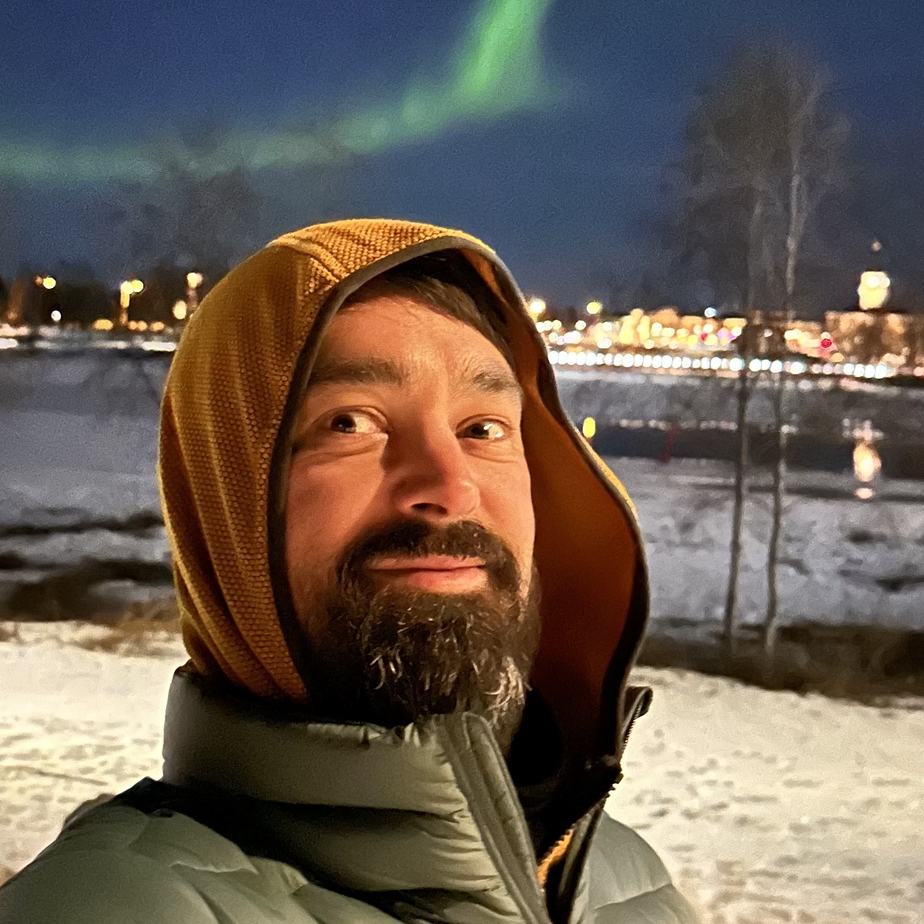 Rob Whitaker wearing a mustard-yellow hood and a grey puffer jacket, photographed outdoors at night in a snowy landscape with the northern lights visible in the sky behind them.